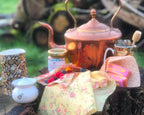 Vintage-style tea set with teapot, cups, and utensils on a floral cloth outdoors.