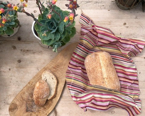 Loaf of bread on a striped cloth with a wooden cutting board and plants on a wooden surface